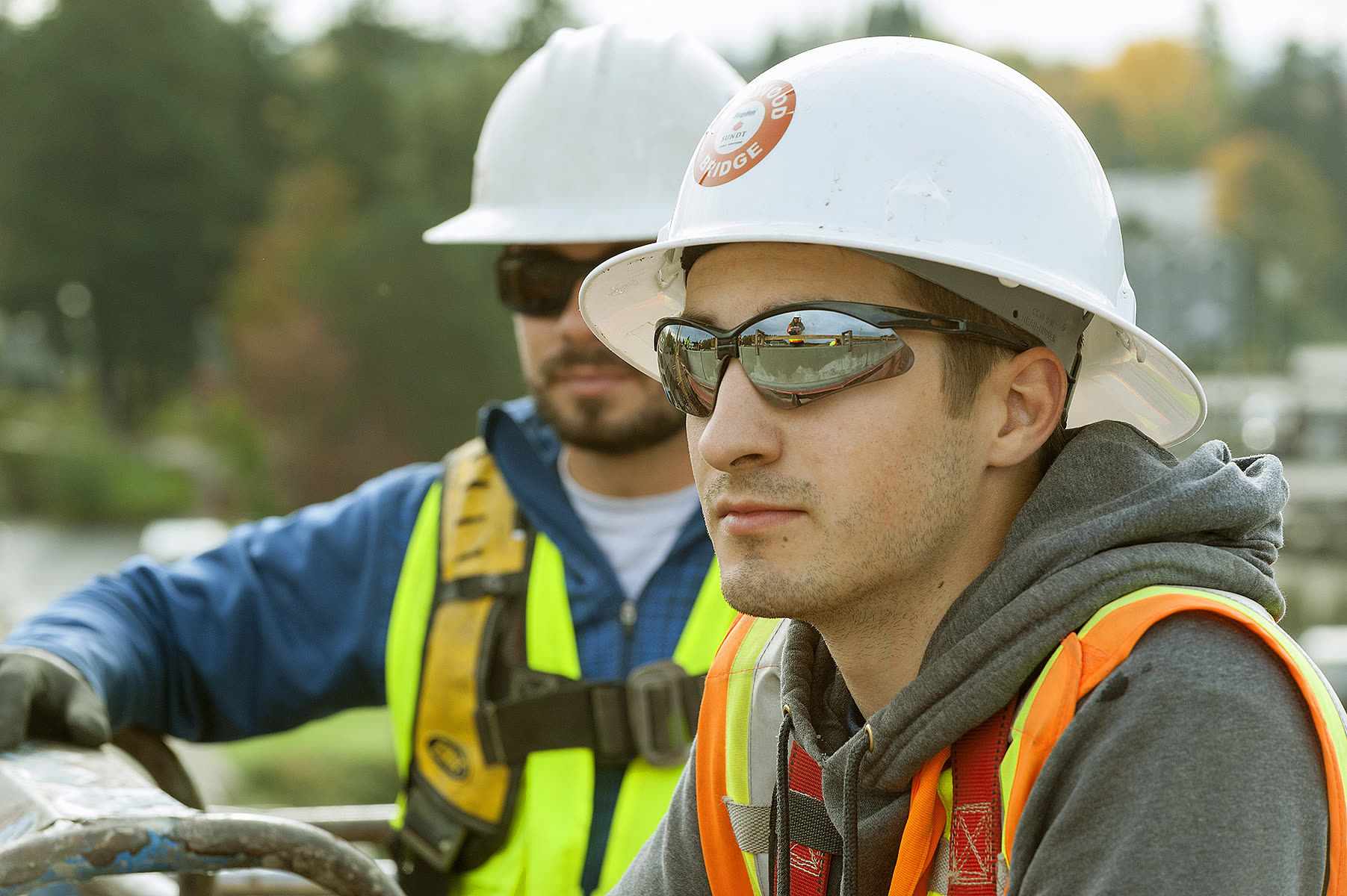 A young apprentice electrician in a white hard hat, safety glasses and hi-vis vest on a construction site, with a co-worker behind him