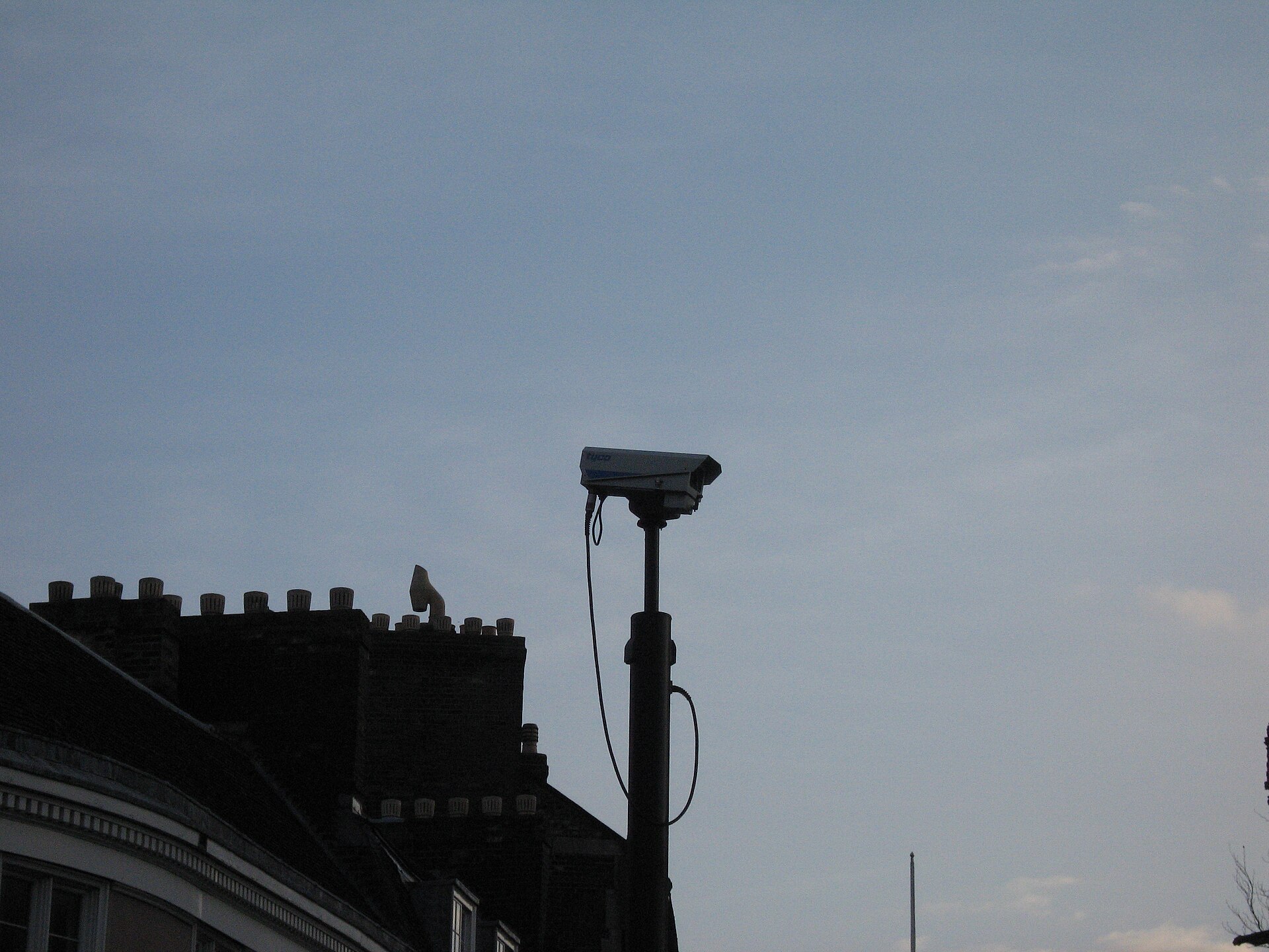 A CCTV camera on a pole silhouetted against a grey sky beside a row of rooftops and chimneys