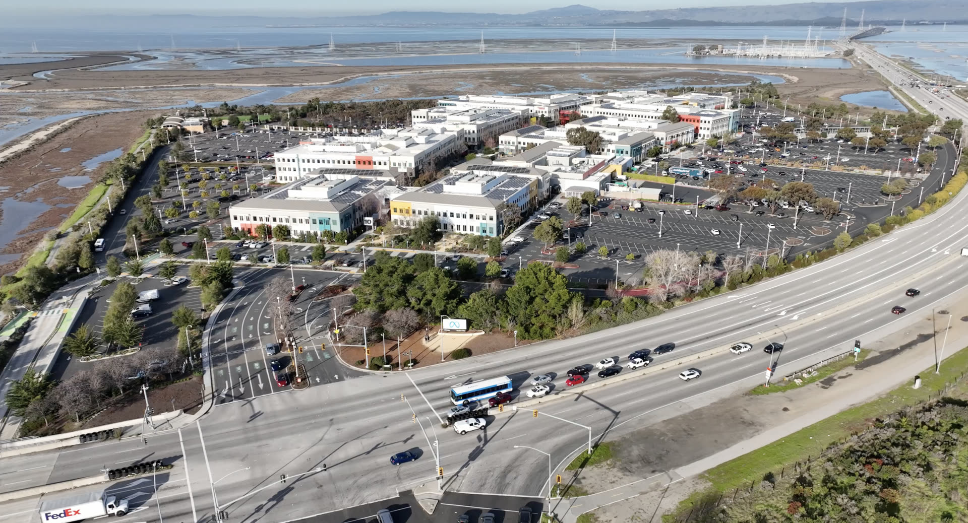 Aerial view of Meta's headquarters campus in Menlo Park, California, beside the bay and a busy road, with the Meta logo sign at the entrance
