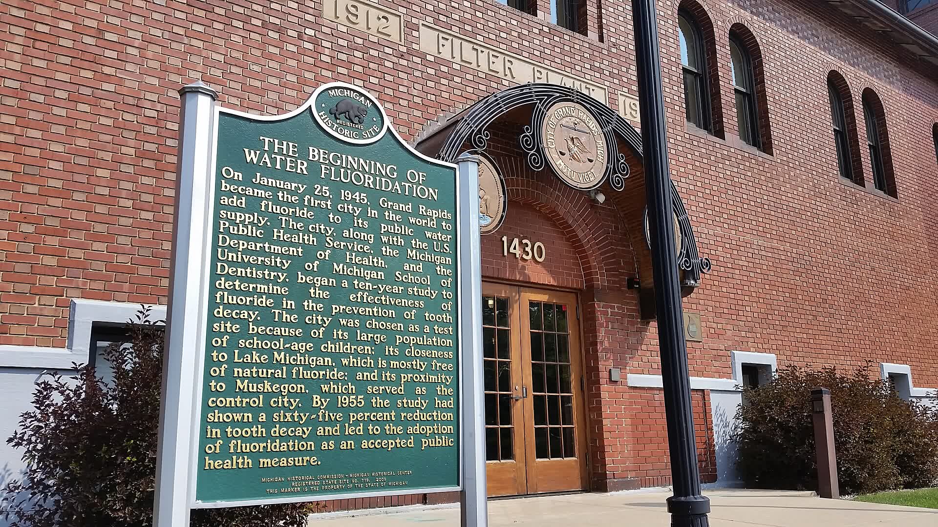 A green historical marker titled The Beginning of Water Fluoridation outside a red brick water filtration plant