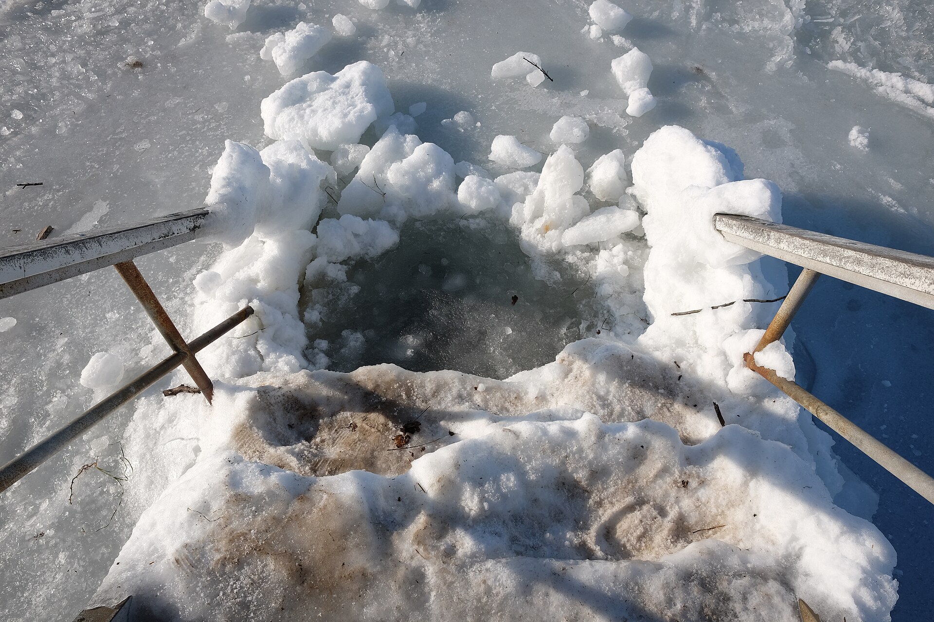 A hole cut into thick sea ice, with metal steps leading down into the dark water, used for winter swimming