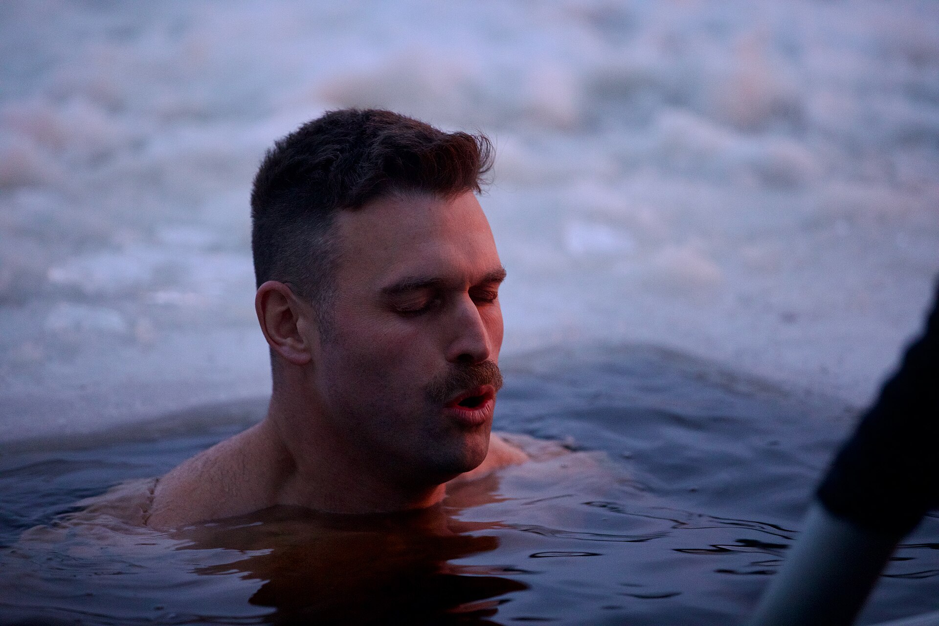 A soldier up to his shoulders in a hole cut in the ice, eyes closed, with snow and ice behind him