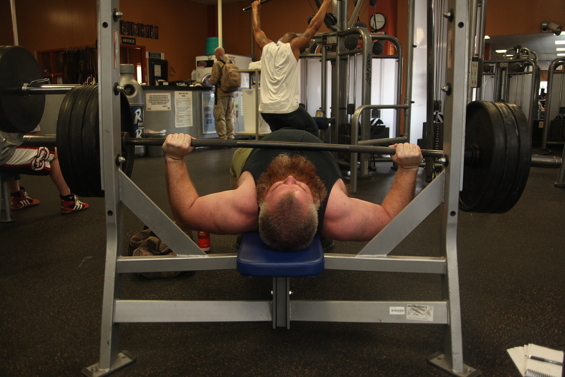 A man lying on a bench in a gym pressing a loaded barbell, watched by a spotter
