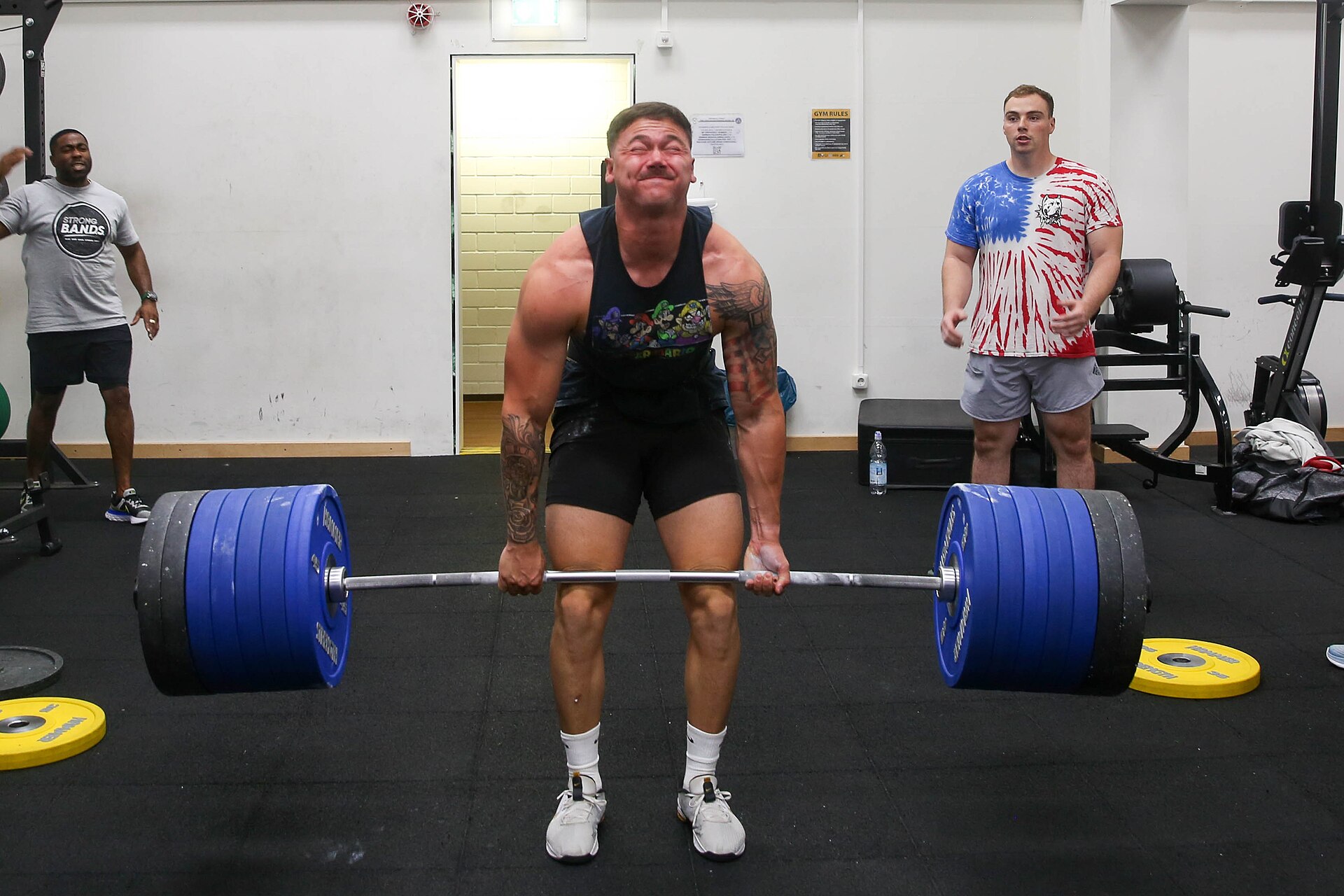 A muscular man in a gym straining as he lifts a heavily loaded barbell off the floor in a deadlift